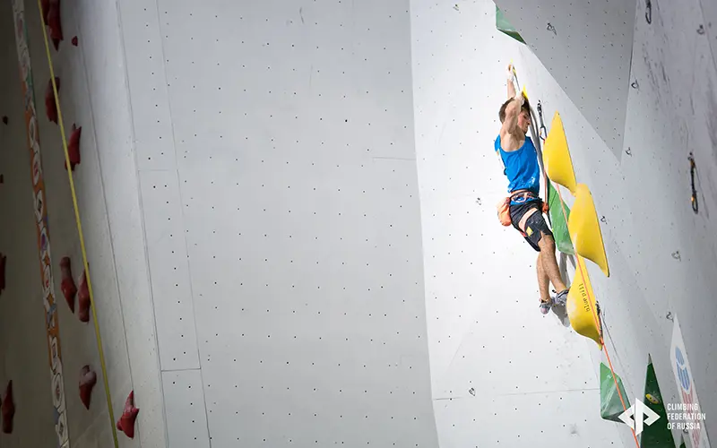 Een klimmer in een blauw pak beklimt een indoor-klimwand en houdt zich vast aan grote gele grepen. De wand is wit en voorzien van geometrische vormen; aan de linkerkant zijn touwen en klimgrepen te zien.